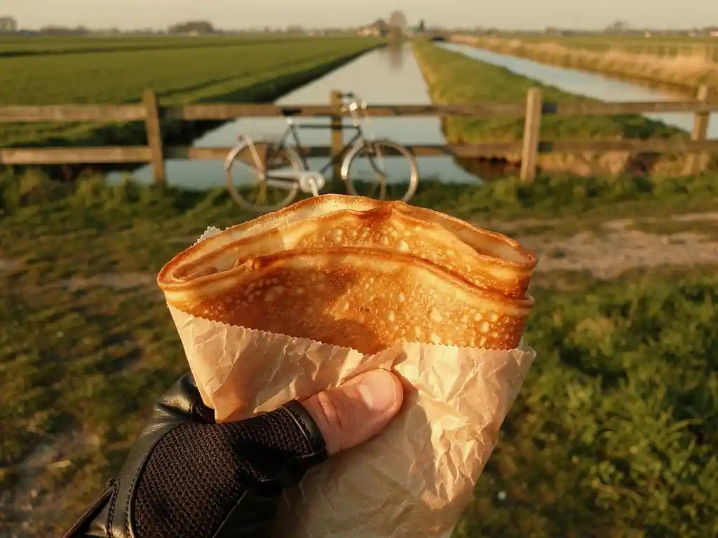 Gouden Dutch pancake in perkamentpapier gehouden door een fietser met handschoenen, met een Hollands polderlandschap op de achtergrond.