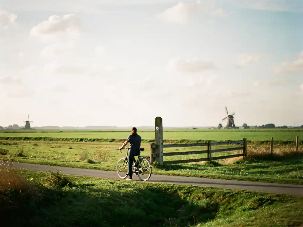 Fietser rustend op smal polderpad in Nederland, fiets tegen houten paal, groene weilanden en windmolens op achtergrond in middaglicht.