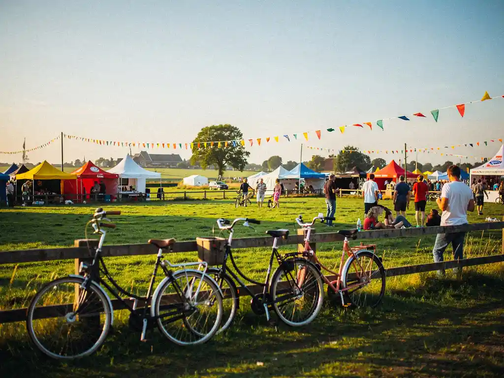 Fietsers stallen hun fietsen bij een houten hek tijdens een zomers buitenfestival op een Nederlands polderlandschap met kleurrijke tenten.